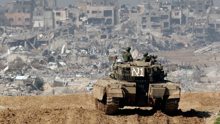 A picture taken from a position in southern Israel along the border with the Gaza Strip on January 19, 2024, shows an Israeli tank rolling along the fence as damaged buildings are see in the Gaza strip amid continuing battles between Israel and the militant group Hamas.