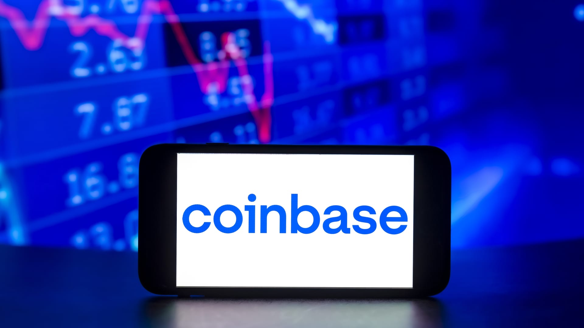 The Coinbase logo is displayed on a mobile phone screen with stock market percentages in the background.